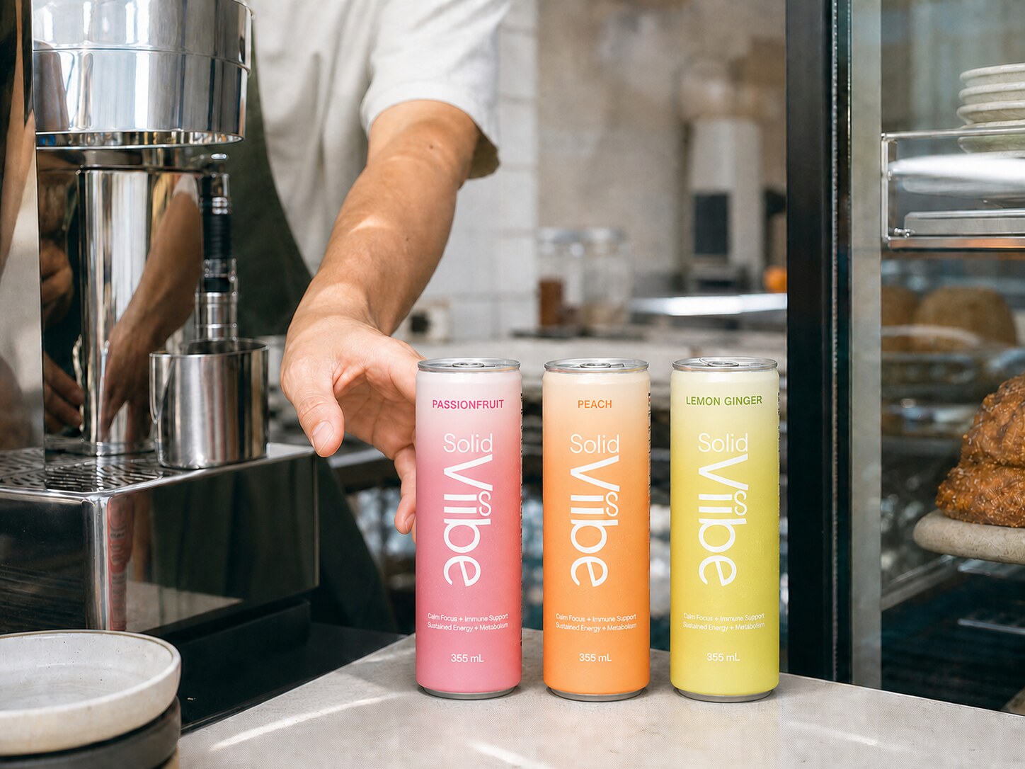 A barista reaching for a can of Passionfruit Solid Viibe from a row of four cans lined up on a cafe counter beside the espresso machine