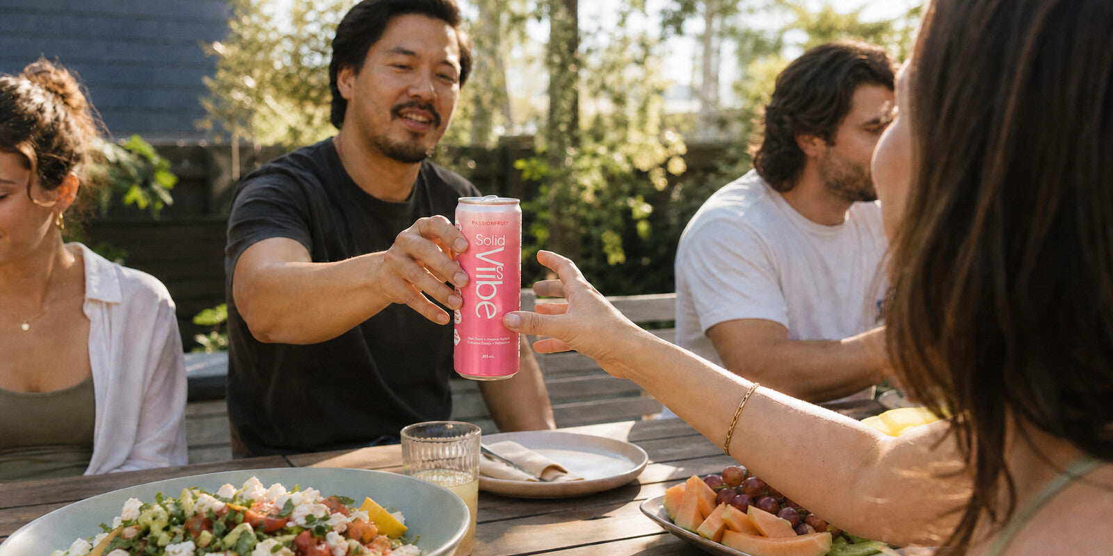 Friends around a backyard table at golden hour, one passing a can of Passionfruit Solid Viibe across a shared meal